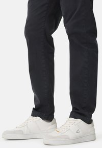 White sneakers with grey accents and textured overlays. Laces are prominent. Worn with black trousers, showing a tapered leg design.