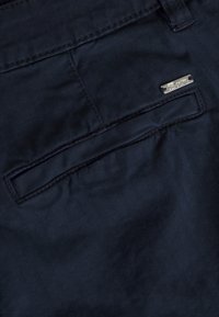 Tessuto blu scuro con una tasca posteriore dotata di un'etichetta metallica rettangolare argento incisa con la scritta "Dart". La trama appare liscia con un motivo sottile.