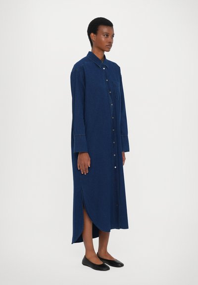 Lang denim skjortekjole i mørkeblå. Har frontknaplukning, krave og sideskår. Stylet med sorte flade sko.