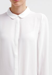 Camisa blanca de manga larga con cierre de botones y una textura suave y lisa. Presenta un cuello clásico y una tela ligeramente fluida.