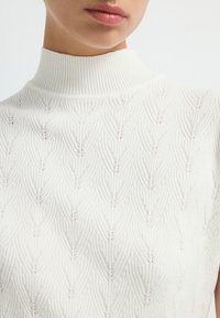 White knitted sleeveless sweater with a high collar, featuring a leaf pattern and ribbed texture. The design is minimal and elegant.