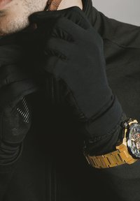 Gants noirs avec des accents texturés, portés sur une veste noire, accompagnés d'une montre dorée avec un bracelet en métal et un cadran audacieux et multicolore.