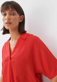 Rote Bluse mit kurzen Ärmeln und V-Ausschnitt, die über eine weiche Textur und eine drapierte Silhouette verfügt. Glattes Material, minimale Akzente.
