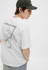 Hvid bomulds t-shirt med en sort kontur grafik af en plante og teksten "JUAN-LES-PINS" på ryggen, med korte ærmer og afslappet pasform.