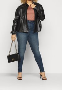 Kvinna som bär en svart läderjacka, lavendelfärgad skjorta, blå tajta jeans, svarta högklackade sandaler och håller en svart handväska med kedjestropp.