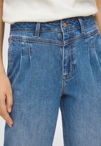 Blue denim pleated jeans with a high waist, featuring a button closure and reinforced stitching along the pockets. Smooth texture.