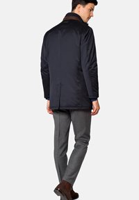 Granatowy ocieplany płaszcz z brązowym kołnierzem i jednym rozporkiem z tyłu, zestawiony z szarymi spodniami i brązowymi botkami za kostkę.