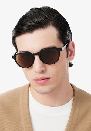 Un homme porte des lunettes de soleil noires aux verres bruns et un cardigan beige sur une chemise blanche, devant un fond uni.