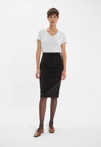 Jupe crayon noire associée à un t-shirt blanc à col en V. Modèle portant des chaussures marron et des collants noirs. Tissu lisse, silhouette ajustée.
