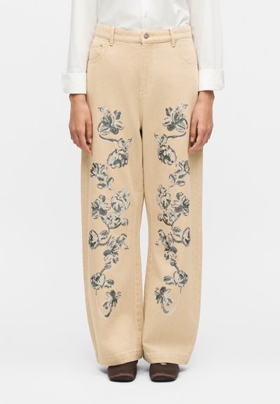 Pantalon beige à jambes larges avec broderie florale noire et blanche sur le devant, porté avec une chemise blanche à manches longues et des chaussures marron.