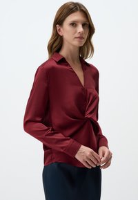 Burgundy satin blouse with a deep V-neck and twisted detail at the waist. Long sleeves with buttoned cuffs, paired with a dark skirt.