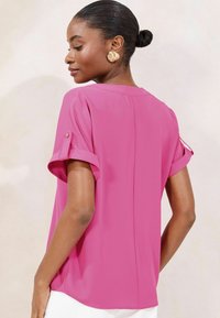Blusa rosa a maniche corte con risvolti, texture setosa e taglio dritto, caratterizzata da un delicato dettaglio di cucitura sul retro.