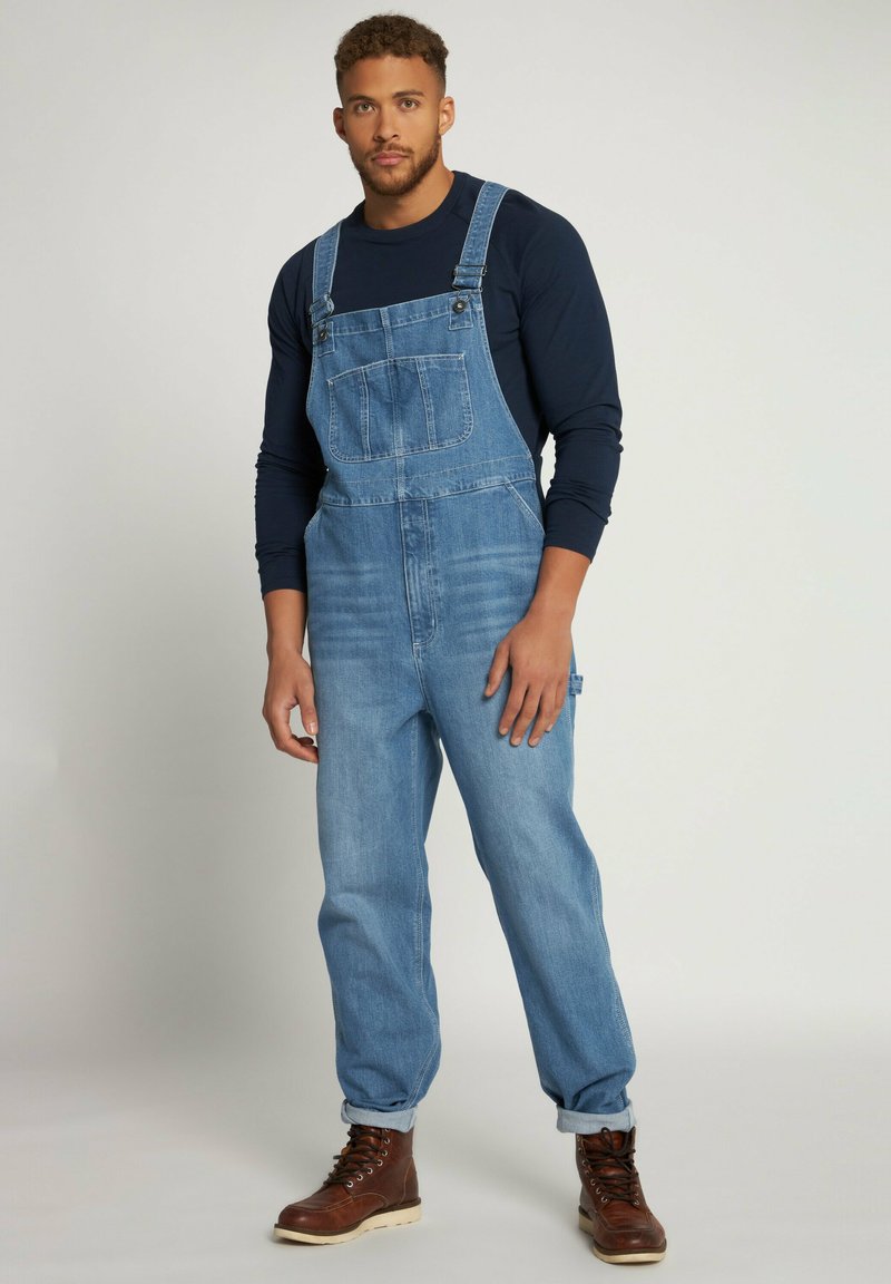 Salopette in denim azzurro chiaro con spalline regolabili, tasca frontale e vestibilità ampia. Indossata con una camicia a maniche lunghe scura e stivali marroni.