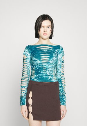 Top de veludo verde-água com mangas longas e recortes horizontais; combinado com uma saia castanha com textura canelada, com detalhes em anel e uma fenda lateral.