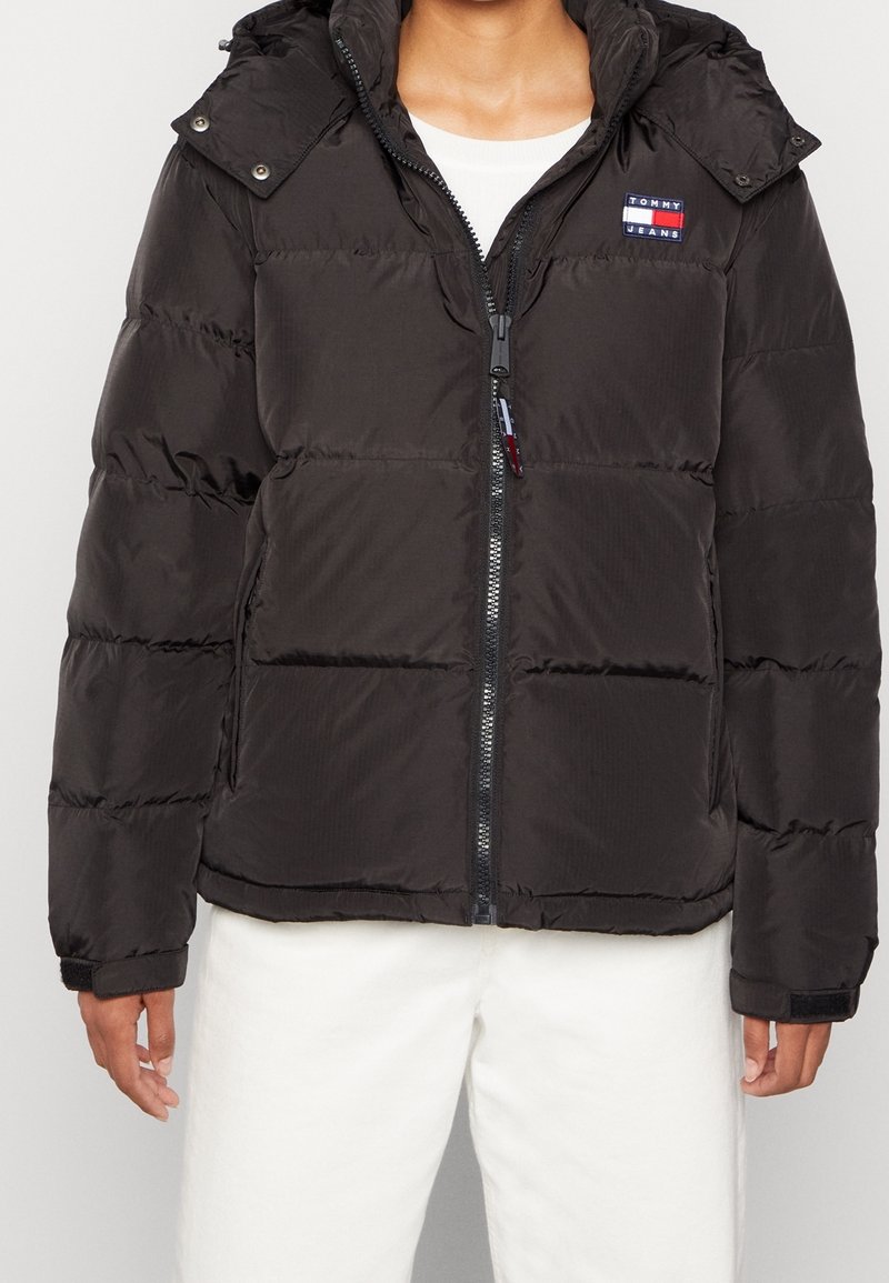 Veste matelassée noire avec fermeture éclair à l'avant, capuche et logo Tommy Jeans sur la poitrine, portée avec un pantalon et une chemise blancs.