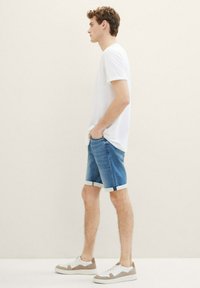 Camiseta blanca de manga corta, pantalones cortos de mezclilla azul con dobladillos enrollados y zapatillas blancas con acentos grises. Perfil de pie contra un fondo neutro.