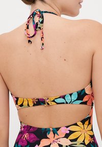 Bloemmotief bikini top met een zwarte achtergrond, voorzien van kleurrijke overgrote bloemen. Verstelbare nekstrik en goudkleurige accenten aan de achterkant.