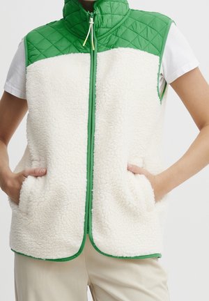 Veste polaire blanche sans manches avec empiècement matelassé vert et bordure portée sur une chemise blanche à manches courtes, mains dans les poches de la veste.