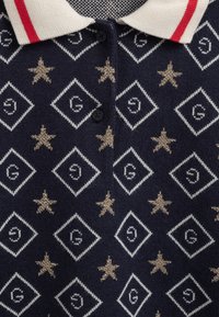 Granatowy sweter polo z teksturowanym wzorem w diamenty i gwiazdki w kolorze złotym i białym. Ma beżowy kołnierzyk z czerwonymi paskami i czarnymi guzikami.