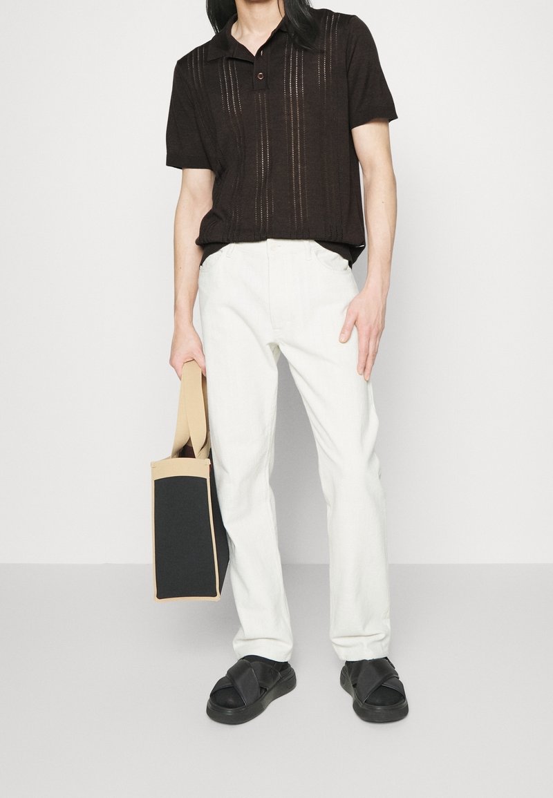 Hnědá, texturovaná polo tričko s límečkem a knoflíkem, spárované s bílými kalhotami rovného střihu. Černé sandály a béžová taška s černým panelem.