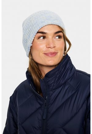 Kvinde iført en lys blå strikket hue og en marineblå quilted jakke, smilende og kiggende let til siden.