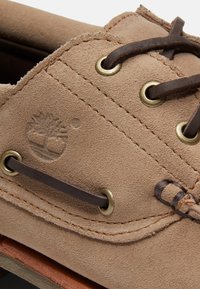 Nærbillede af tan ruskindssko med brune læder snørebånd trukket gennem messingnitter og et præget trælogo på siden.