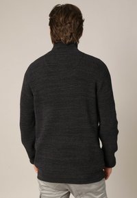 Man met gemiddeld lang bruin haar, gekleed in een donkergrijze gestructureerde gebreide trui en lichtgrijze broek, die met zijn rug naar de camera staat.