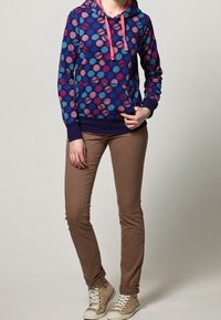 Sweat à capuche violet avec un motif de points multicolores, cordons de serrage et poche avant. Associé à un pantalon ajusté marron clair et des baskets beiges.