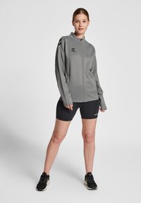 Grå zip-up træningsjakke med lange ærmer, glat tekstur, logo på brystet, parret med sorte cykelsort og sorte sneakers.