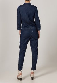 Mörk denim overall med lång ärm, avsmalnande ben och framfickor. Har en figurnära midja och knappryssning på ärmarna. Vita plattformshälar.