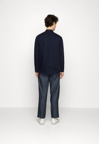 Camisa azul marinho com colarinho pontudo, mangas longas e pregas nas costas; combinada com calças de ganga escuras de perna reta e ténis brancos.