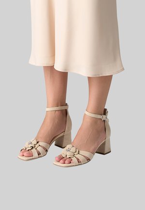 Beige block heel sandals with ankle straps and decorative flower accents worn with a cream-colored midi skirt.