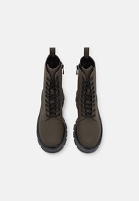 Bottes de cheville vertes avec une finition lisse imitant le cuir, bout rond, semelle en caoutchouc noire et lacets noirs ; elles possèdent une fermeture éclair sur le côté et une languette.