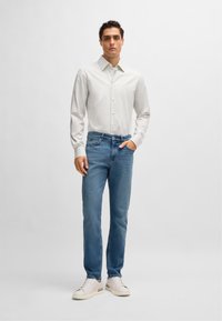 Striped white and light gray long-sleeve button-up shirt, blue denim jeans, and beige sneakers. Model stands with one hand in pocket.