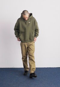 Olīvu zaļš kapuču krekls ar logo, priekšējo kabatu un auklītēm; kombinēts ar bēšām biksēm un melniem apaviem. Stāv uz zilā paklāja, skatoties uz leju.