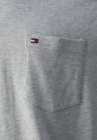 Šedé tričko s náprsní kapsou. Na kapse je vyšitá malá logo v červené, bílé a modré barvě.