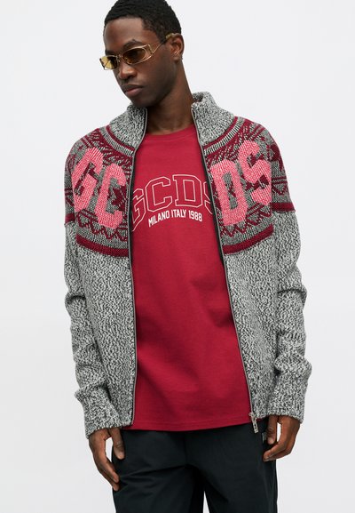 Grijze gebreide jas met rode patroonelementen op de schouders, gedragen over een rood shirt met witte tekst. Model met zonnebril poseert.