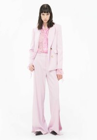 Completo giacca rosa chiaro con blazer strutturato e pantaloni a gamba larga. Blusa rosa trasparente sottostante con un motivo sottile. Calzature nere con accenti.