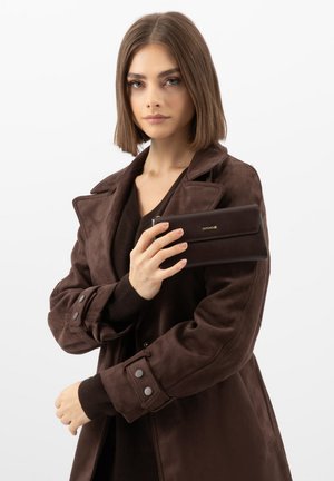 Jonge vrouw met steil bruin haar draagt een donkerbruine jas en houdt een bijpassende bruine portemonnee met gouden logo vast tegen een witte achtergrond.