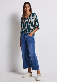 Blouse à manches longues et décolleté en V avec un motif abstrait en teal et crème, associée à un jean en denim bleu à jambes larges et des baskets blanches.