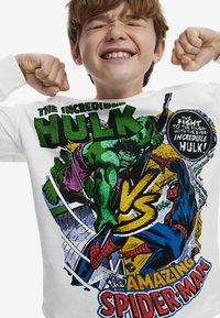 Balta ilgomis rankovėmis marškinėliai su spalvingais komiksų stiliaus grafikos piešiniais apie Hulką ir Žmogų vorą, su ryškiu tekstu, skatinančiu jų konkurenciją.