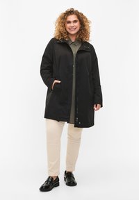 Black waterproof jacket with a high collar, zip closure, and two side pockets, worn over a light green shirt and beige pants.