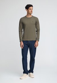 Olivgrön långärmad tröja med rund hals, kombinerad med mörkblå jeans och beige sneakers. Har en liten logga på bröstet.