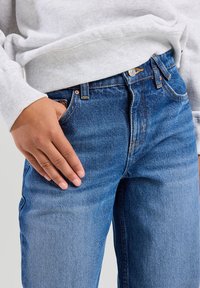 Siniset farkut, joissa on keskitason pesu, suora leikkaus, näkyvät ompeleet ja viisi taskua. Vyötäröllä vaalea harmaa collegepaita.