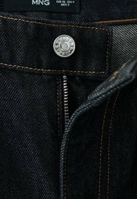 Vaqueros de mezclilla negra con costuras naranjas, cremallera metálica y cierre con botón plateado. El interior tiene un forro de tela texturizada.