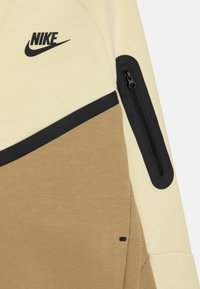 Oblečenie Nike s béžovým a hnedým farebným blokovým dizajnom, s čiernou zipsovou vreckom na rukáve a logom na hrudi. Textúrovaná tkanina.