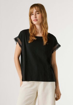 Jonge vrouw met steil rood haar, gekleed in een zwart kortärmelig shirt met kanten rand en witte broek, tegen een effen achtergrond.