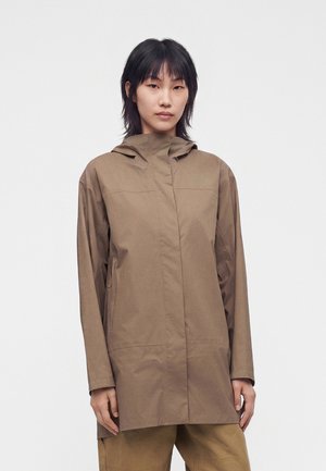 Frau mit kurzen schwarzen Haaren, die eine braune Kapuzenjacke und passende braune Hose trägt, steht vor einem einfarbigen hellen Hintergrund.