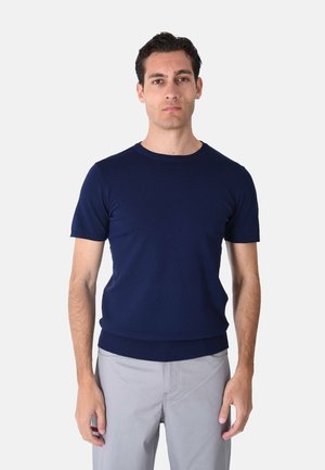 Uomo che indossa una maglietta a maniche corte calata blu scuro e pantaloni grigio chiaro, in piedi davanti a uno sfondo bianco semplice.