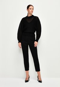 Svart stickad tröja med genomskinlig randig struktur och vida ärmar, kombinerad med tighta svarta byxor och högklackade skor, vilket skapar en monokrom look.